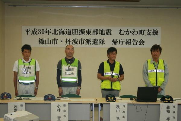 平成30年度北海道胆振東部地震むかわ町支援の帰庁報告会で反射ベストを着用した、1次派遣の河野主事、小林主査、篠山市職員2人が立って挨拶をしている写真