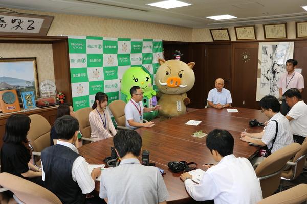 応接室に着席する関係者と、まるいのとまめりんの横で話す男性の写真