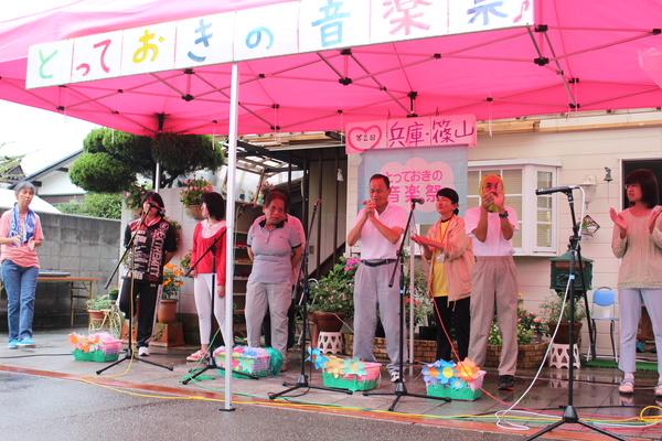 とっておきの音楽祭と紙が貼られたピンクのテントの下で8名ほどの男女がマイクの前に立っている写真