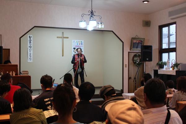 十字架の飾ってあるステージの上で男性が大きな笛を吹いていて、10名ほどの人が聞いている写真