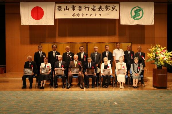 篠山市善行者表彰式と書かれていいる横断幕と金屏風の前で、受賞者8名と関係者が一緒に写っている記念撮影の写真