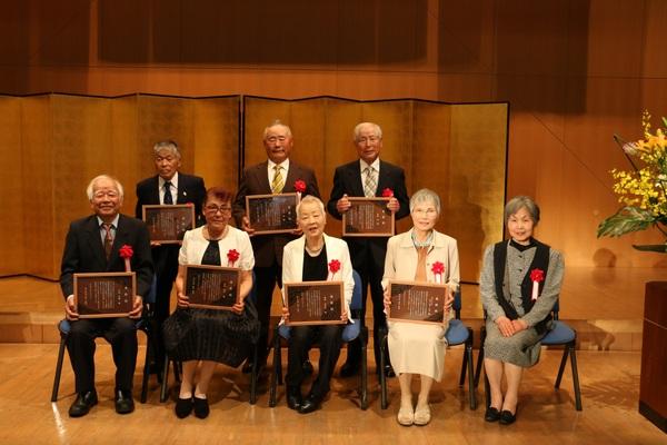 金屏風の前で、受賞者8名が額に入った表彰状を持って記念撮影をしている写真