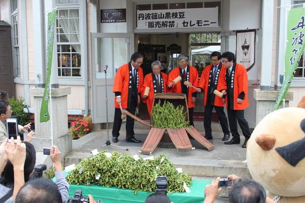 オレンジの法被を着た男性5人が箱を金づちで割り沢山の黒枝豆が出てきている写真
