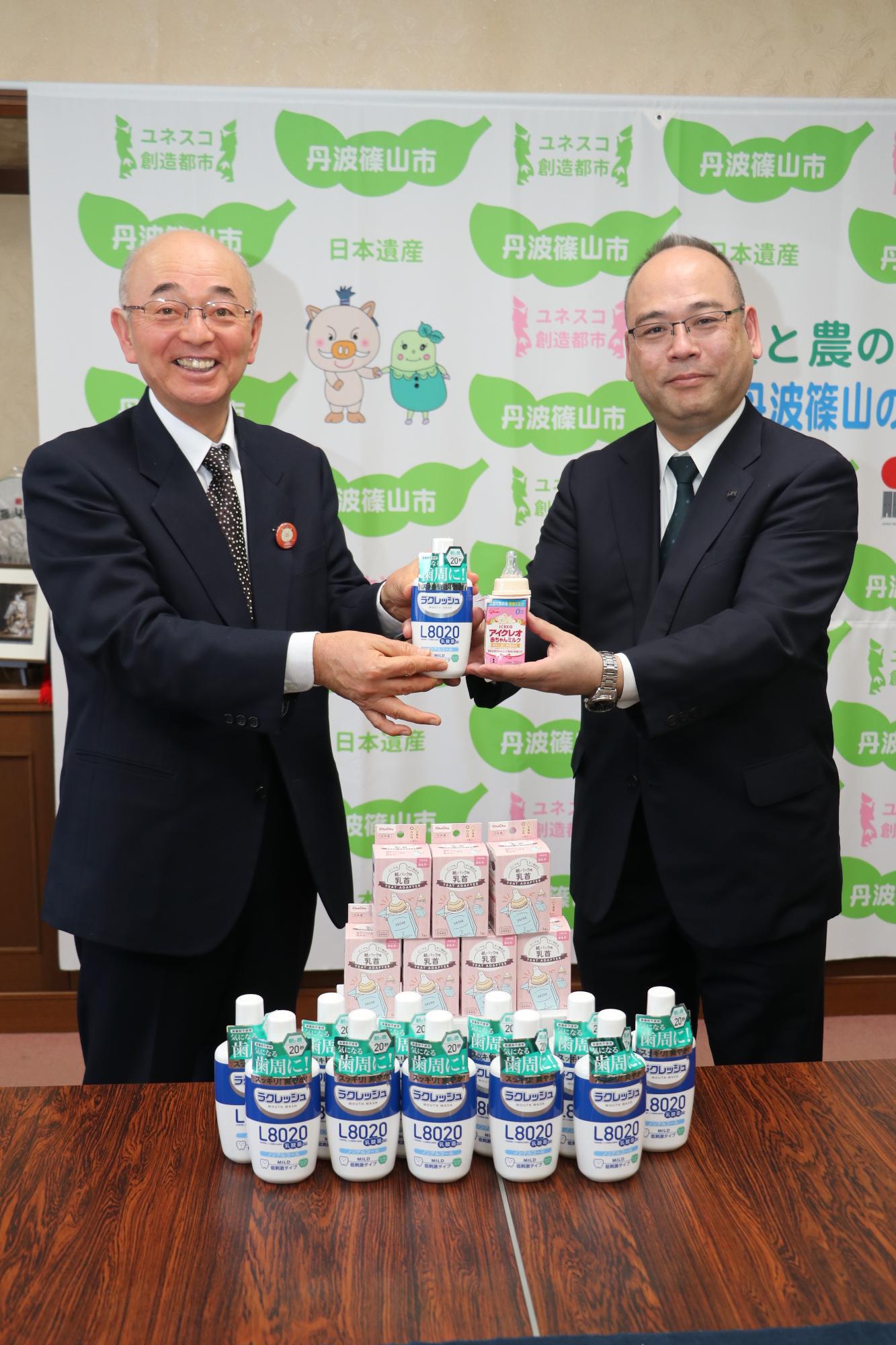 ジェクス株式会社から寄贈して頂いた洗口液と紙パック用乳首を机に並べ、市長が洗口液を、梶川社長が紙パック用乳首を持って写っている写真