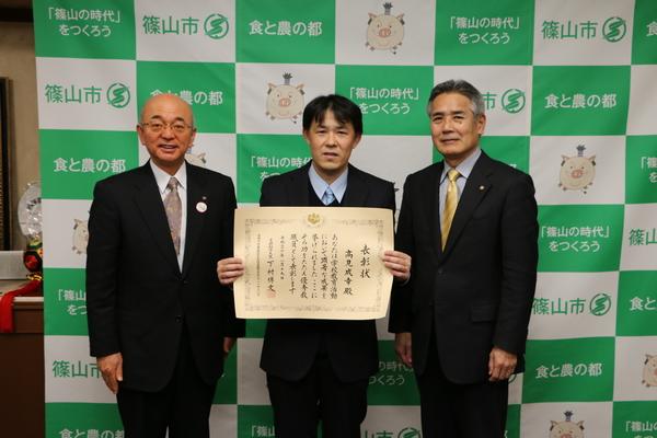高見先生が表賞状を胸の前に持って見せ、市長と金色のネクタイを締めた男性と3名で写っている写真