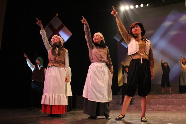 エプロン姿の女性役2人と男性役1人が右手を上げて遠くをみている同じ仕草をその後方の出演者の人達もしている写真