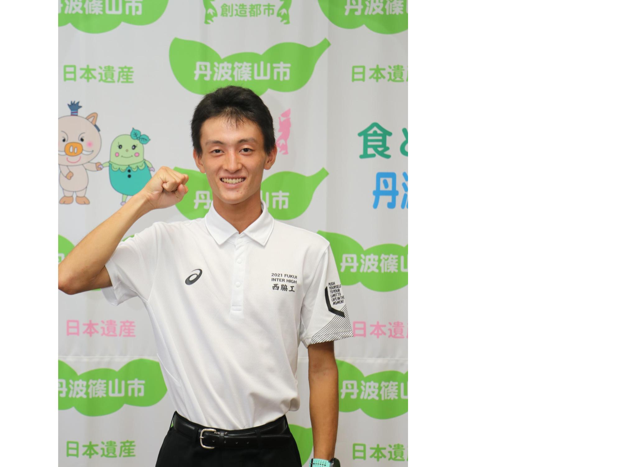 陸上部のポロシャツを着た男子高校生が、タペストリーの前でガッツポーズをとっている