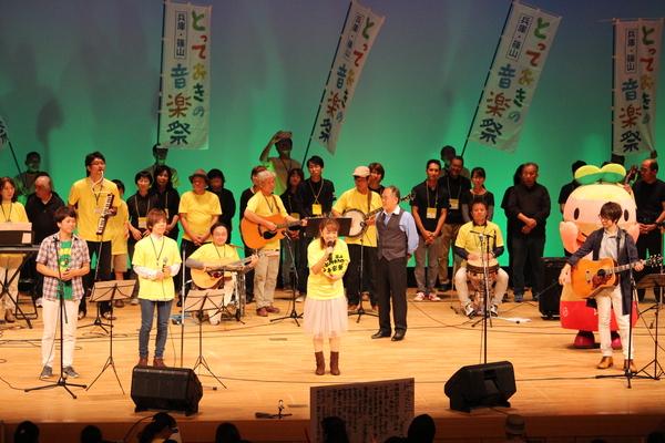 ギターや太鼓を持った男女がステージに立っていて、中央では黄色いTシャツの女性がマイクで歌っている写真