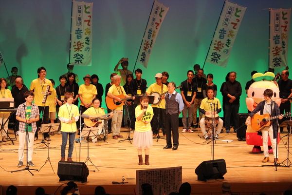 ギターや太鼓を持った男女がステージに立っていて、中央では黄色いTシャツの女性がマイクで歌っている写真