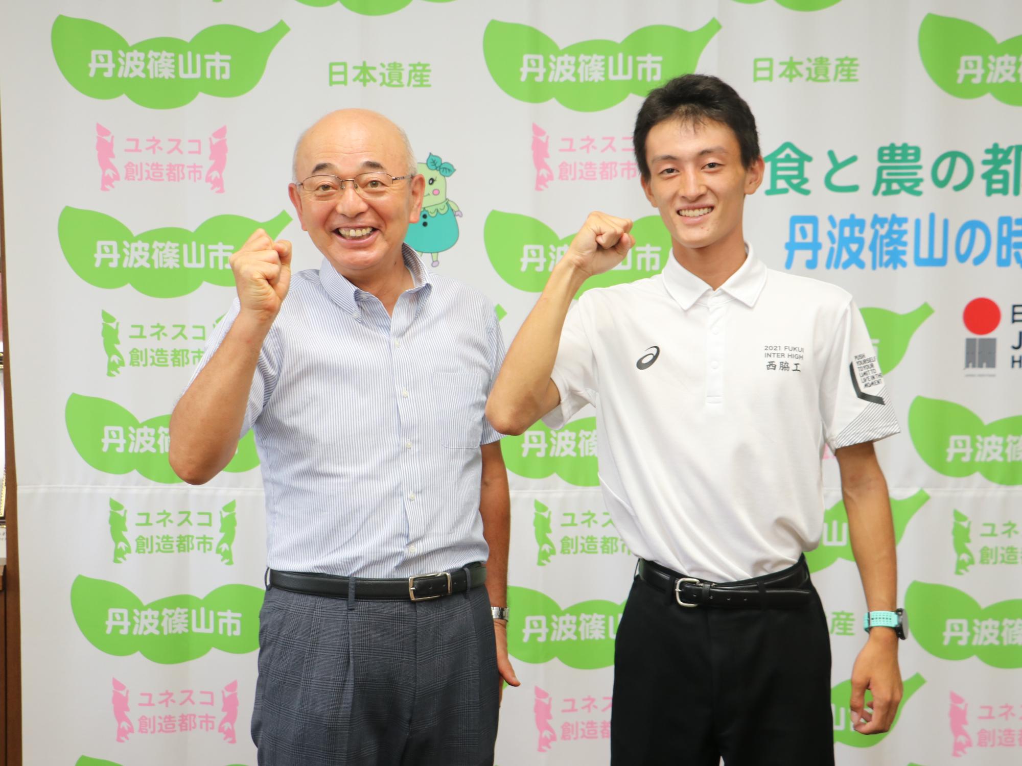 陸上部のポロシャツを着た男子高校生と市長が、タペストリーの前でガッツポーズをしている