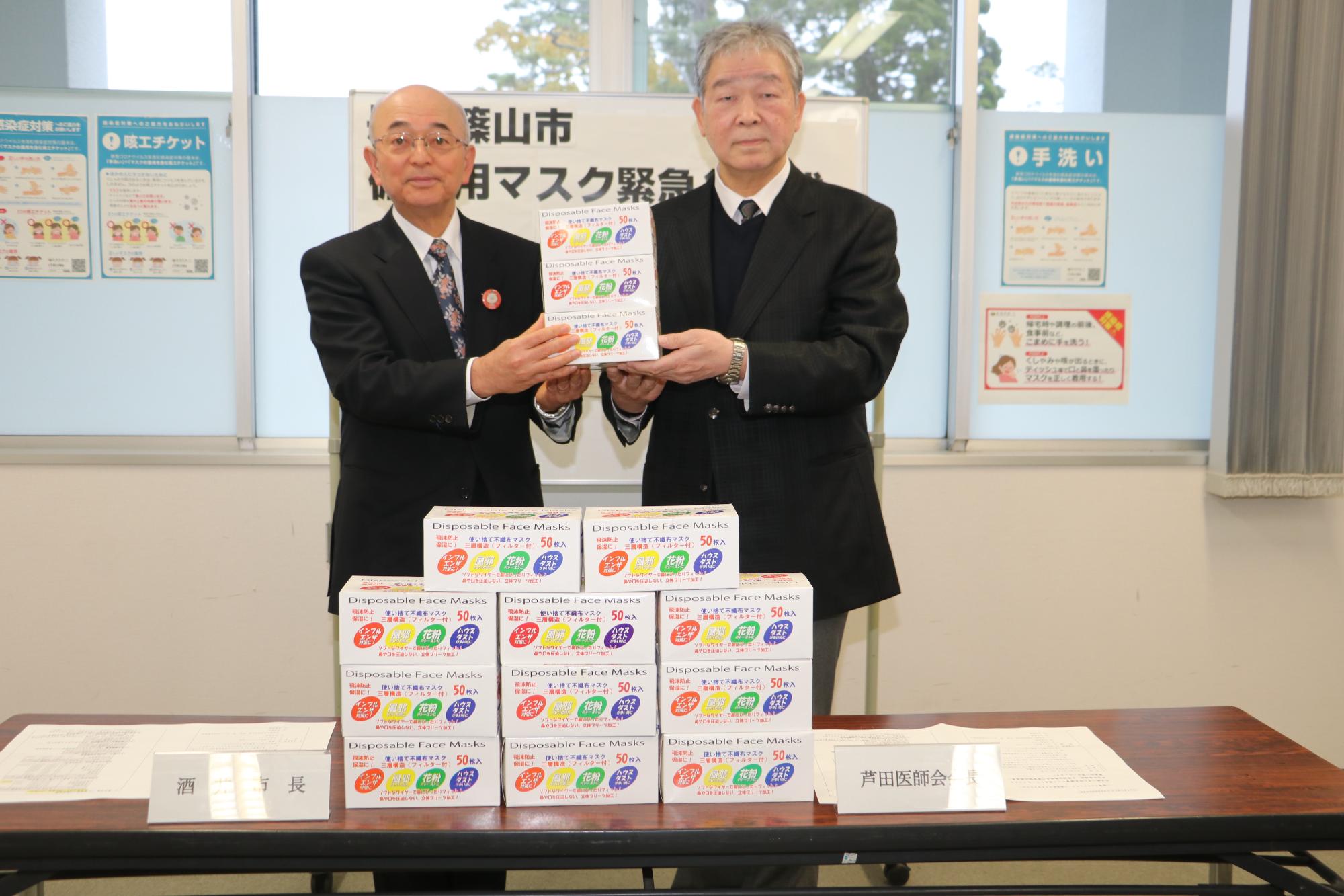 机に市から寄贈するマスクの箱が並べられ、医師会会長の芦田先生と酒井市長が立って隣に並び、マスクの箱を一緒に持っている写真