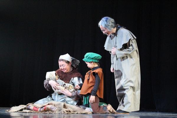 座っている女性が赤ちゃんを抱き、その赤ちゃんの様子を見ている子供と老婆役の人の写真