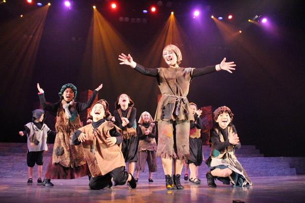 茶色の衣装を着て両手を広げている女性が立っており、その周りの子供たちが笑顔で手を叩いている写真