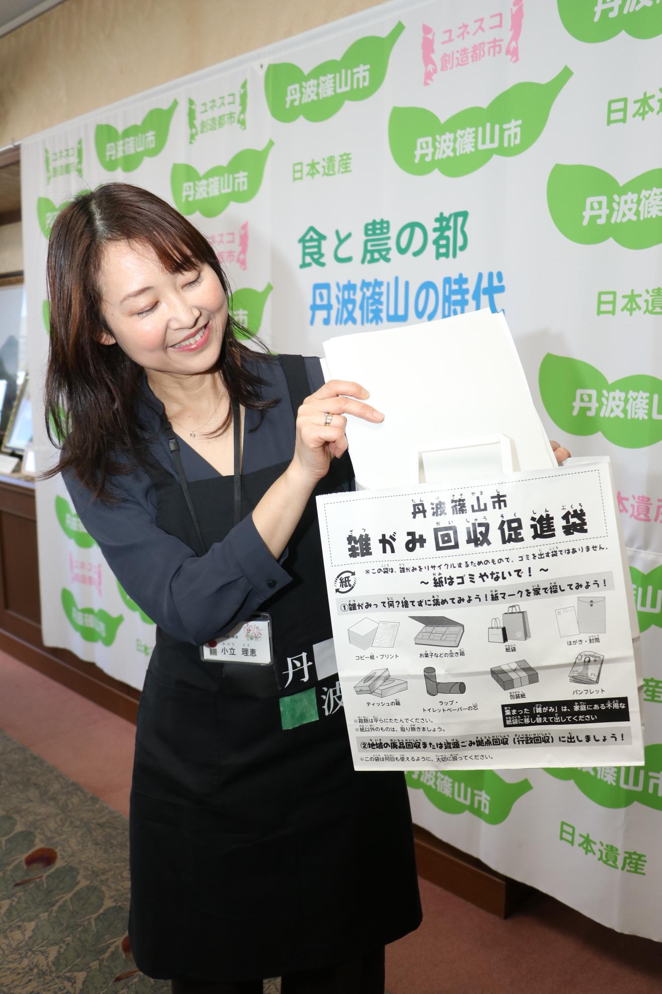 黒色のエプロンを着た市の職員が雑がみ回収促進袋を持ち、雑がみをその中にいれている写真