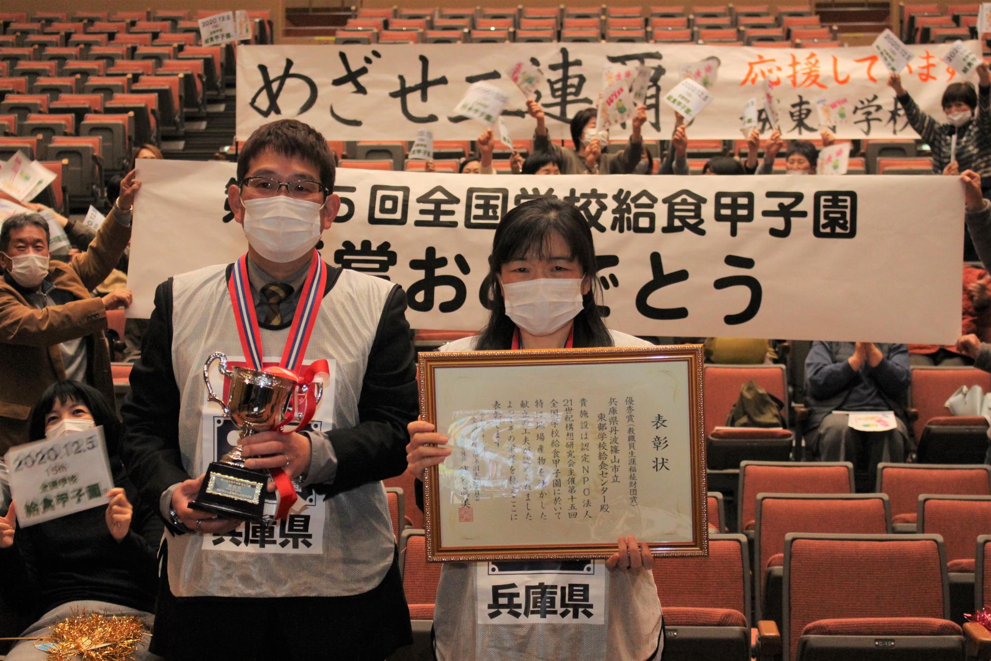 兵庫県と書かれた白色のビブスをしている職員二人が、トロフィーと額に入った表彰状をそれぞれ手に持っている。後ろには「受賞おめでとう」「めざせ「2連覇」という横断幕が掲げてある。