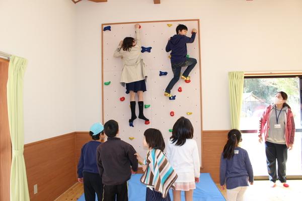 施設内のクライミングウォールを児童2名が登りそれを児童クラブの先生や子供たちが見守っている写真