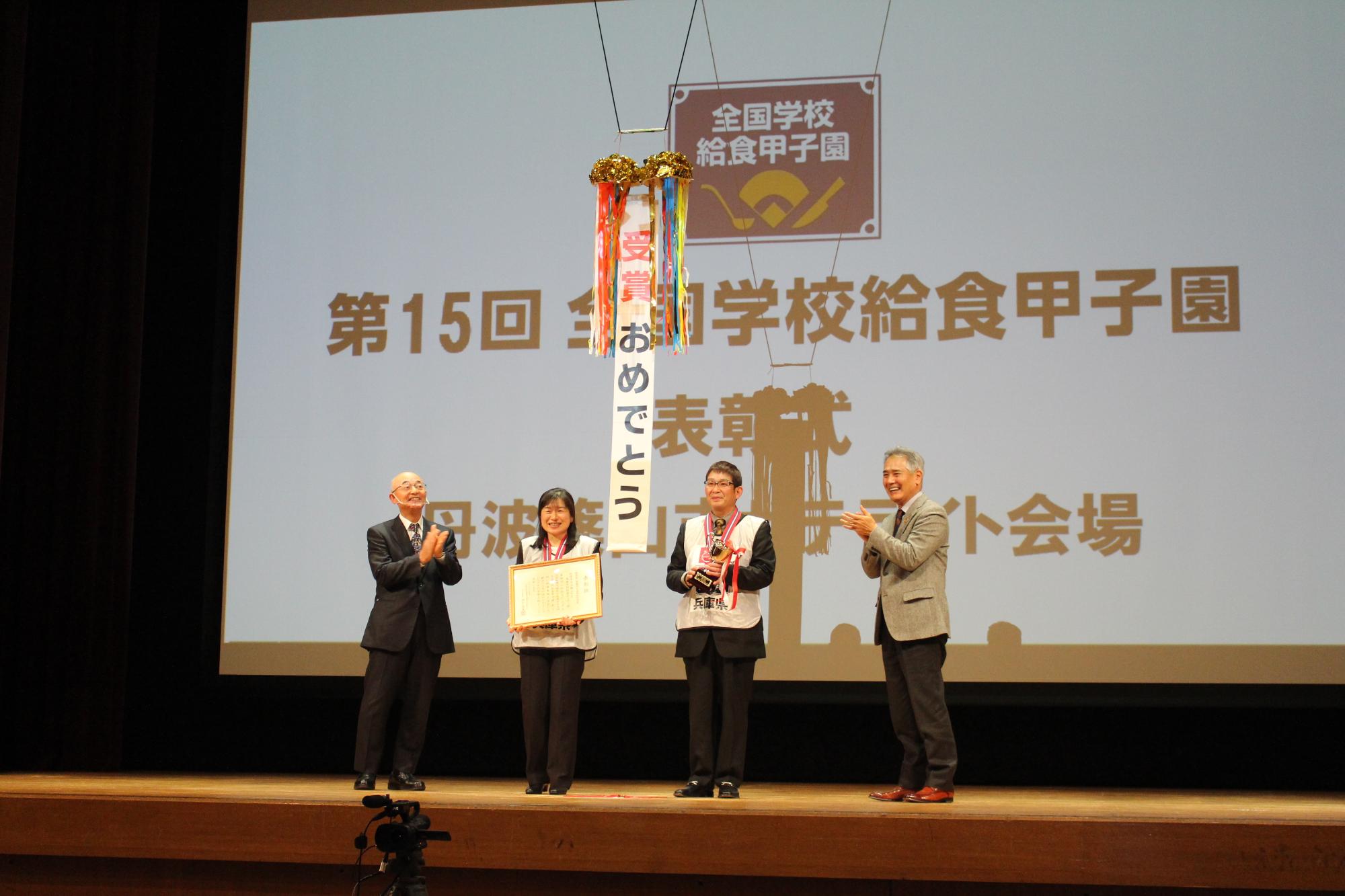 舞台頭上に準備されたくす玉を割り、「受賞おめでとう」という文字。舞台には、出場した職員二人が立っており、市長、教育長が拍手している。