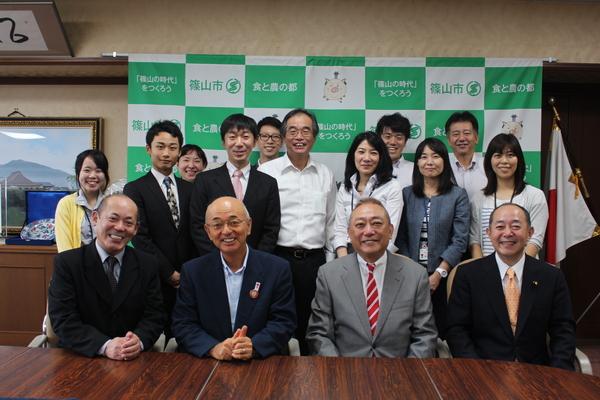 市長と渋谷さん、スーツを着た男性2人が前に座っていて、その後ろに10人ほどの関係者が並んでいる集合写真