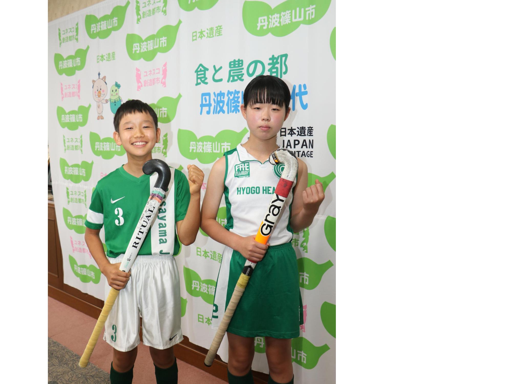 ホッケーのユニフォームを着て、スティックを手に持った男の子と女の子がガッツポーズをしている。