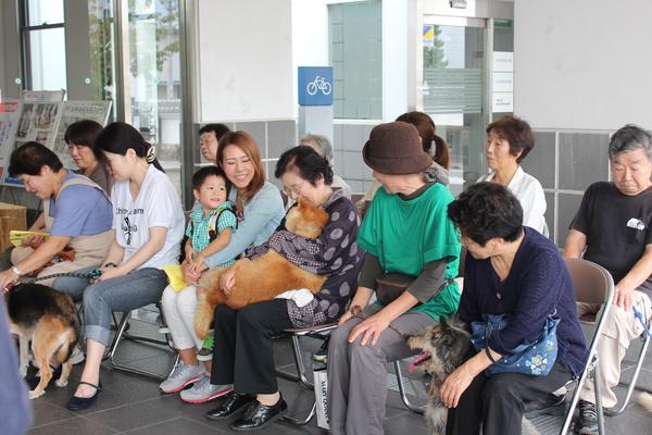 水玉模様の洋服を着て眼鏡をかけている女性が椅子に座って長寿犬を抱っこしている様子を笑顔で見ている親子らの写真