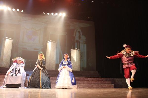 白い衣装を着て椅子に座っている男性の横にドレスを着た女性2名が立っており、右前方には両手を広げ左足をあげている男性の写真