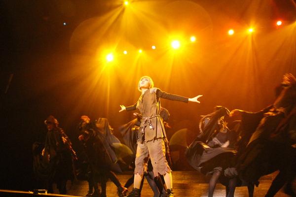 茶色の衣装を着て両手を広げている女性と黒の衣装を着た出演者に黄色のライトが当たっている写真