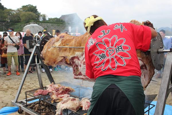 針金でさされた肉のかたまりを赤い服を着た男性が焼いている写真
