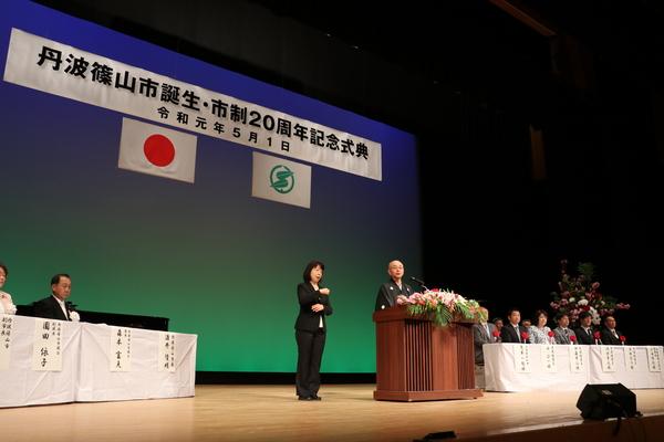 波篠山市誕生祭で市長が話をしていて、横で女性が手話をしていて、来賓者が座っている写真