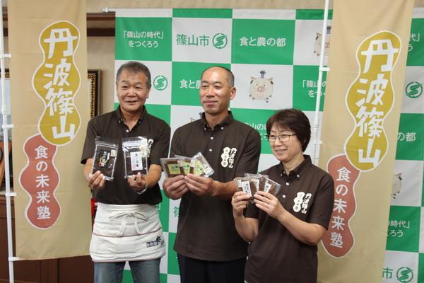 新発売された「黒豆ふりかけ」の商品を両手に持って男性2名と女性1名が笑顔で記者会見を行っている写真