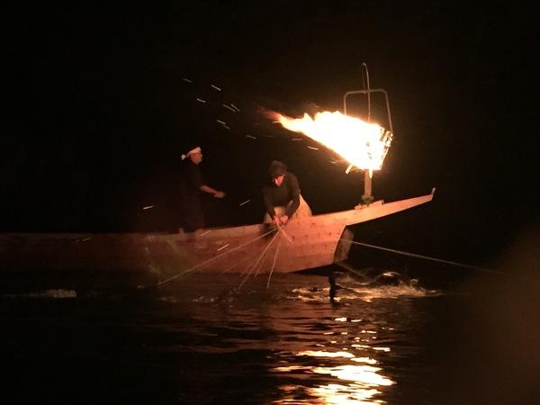 暗闇の中、ボートで、火を焚き、男性が鵜飼いをしている写真