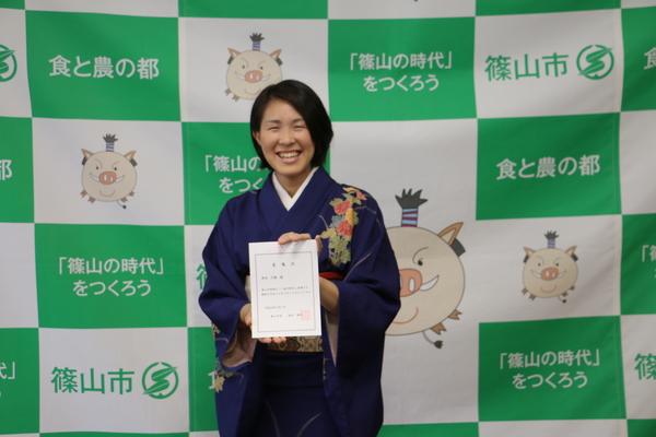 紫色の着物姿の地域おこし協力隊の岸田さんが委託状を両手に持った笑顔の写真
