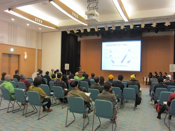 会場の前方のスクリーンに映像を写して発表が行われており、椅子に座って真剣に聞いている出席者の後方から写している写真