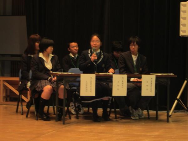 制服を着ている学生3名が机に座っており、真ん中の女子生徒がマイクを持って話をしている様子の写真