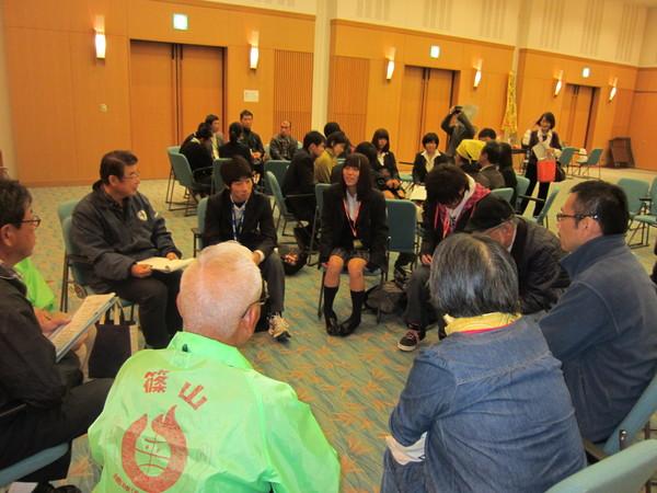 高校生と参加者がグループに分かれて円になって座っており、女子高校生が発言しているのを回りの参加者が興味深く聞いている様子の写真