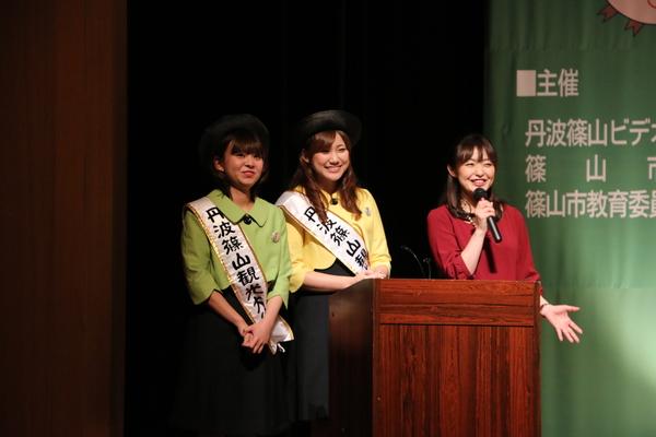 演台の前にマイクを持って話をしている司会の熊谷さんと「丹波篠山観光大使」と書かれているタスキを掛けている黄緑色の上着と来ている女性と黄色い上着を着ている観光大使の女性が写っている写真