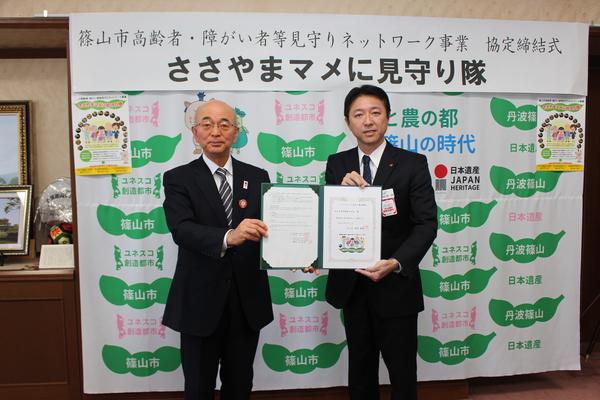 市長とスーツを来た男性が篠山市と書かれた壁の前で協定書を持っている写真