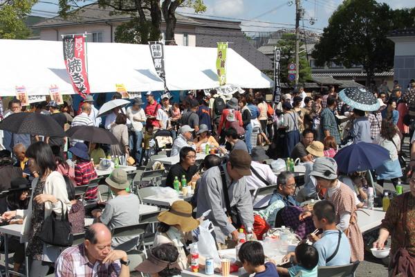 秋晴れの下、篠山味まつりのイートインスペースで多くの人が席に座り食事をしたり、食べ物が販売されているテントの前に多くの人が並んでいる様子の写真