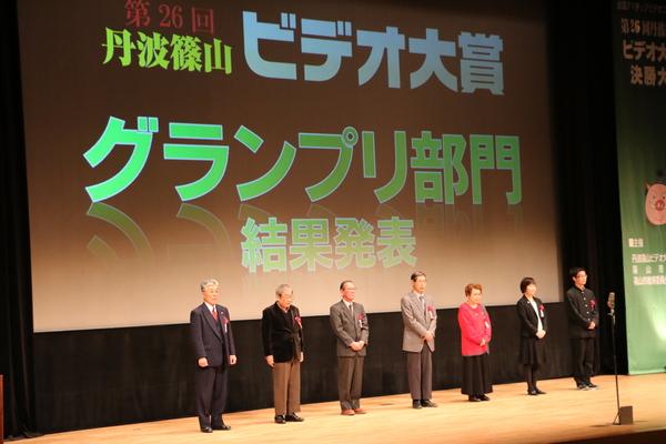 舞台のスクリーンに「第26回 丹波篠山ビデオ大賞 グランプリ部門 結果発送」と出ており、舞台にはノミネートされた方7名が並んで立っている様子の写真
