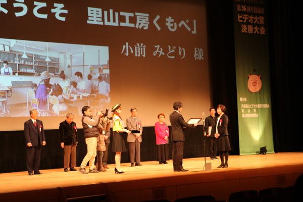 表彰式で中央の受賞者の女性が賞状を読み上げてもらっている様子の写真