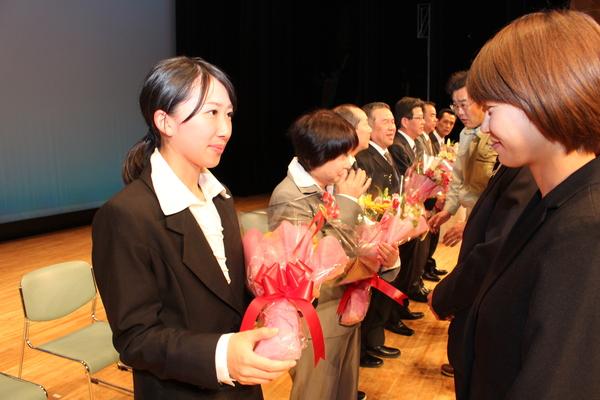 花束を持ち横一列に並ぶ退職者の前に並ぶ、男女の写真