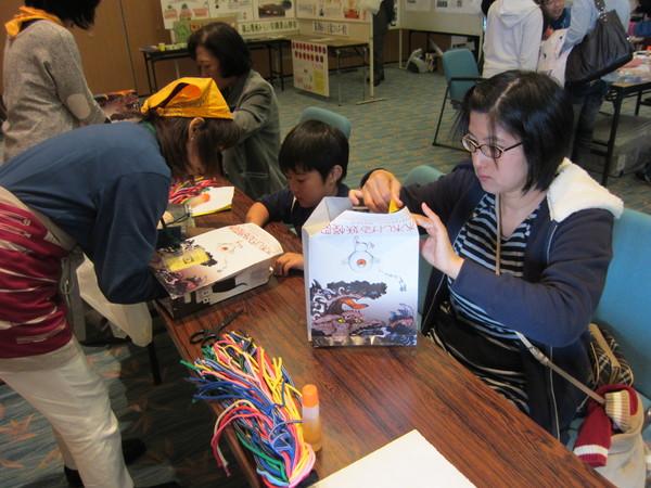 男の子とお母さんが水木茂妖怪図鑑の目玉おやじのイラストが描かれている紙を使って箱のような物を製作をしている様子の写真