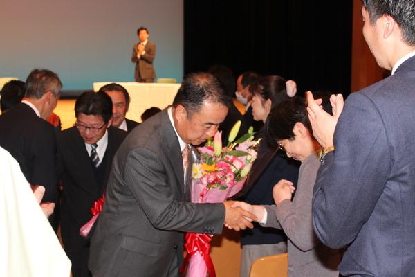 花束を持つ男性退職者が、女性の職員と握手をし、深々と頭を下げている写真