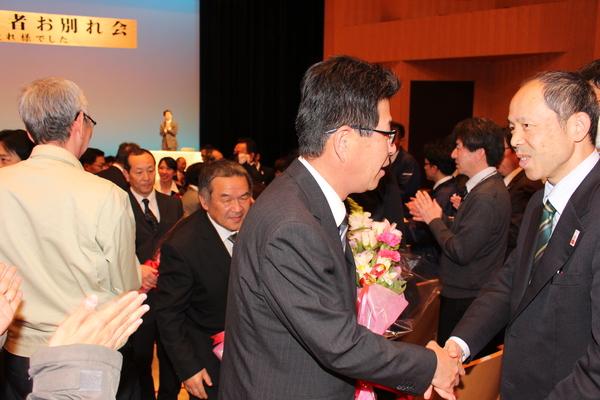 花束を持った退職者が、拍手で見送られている写真