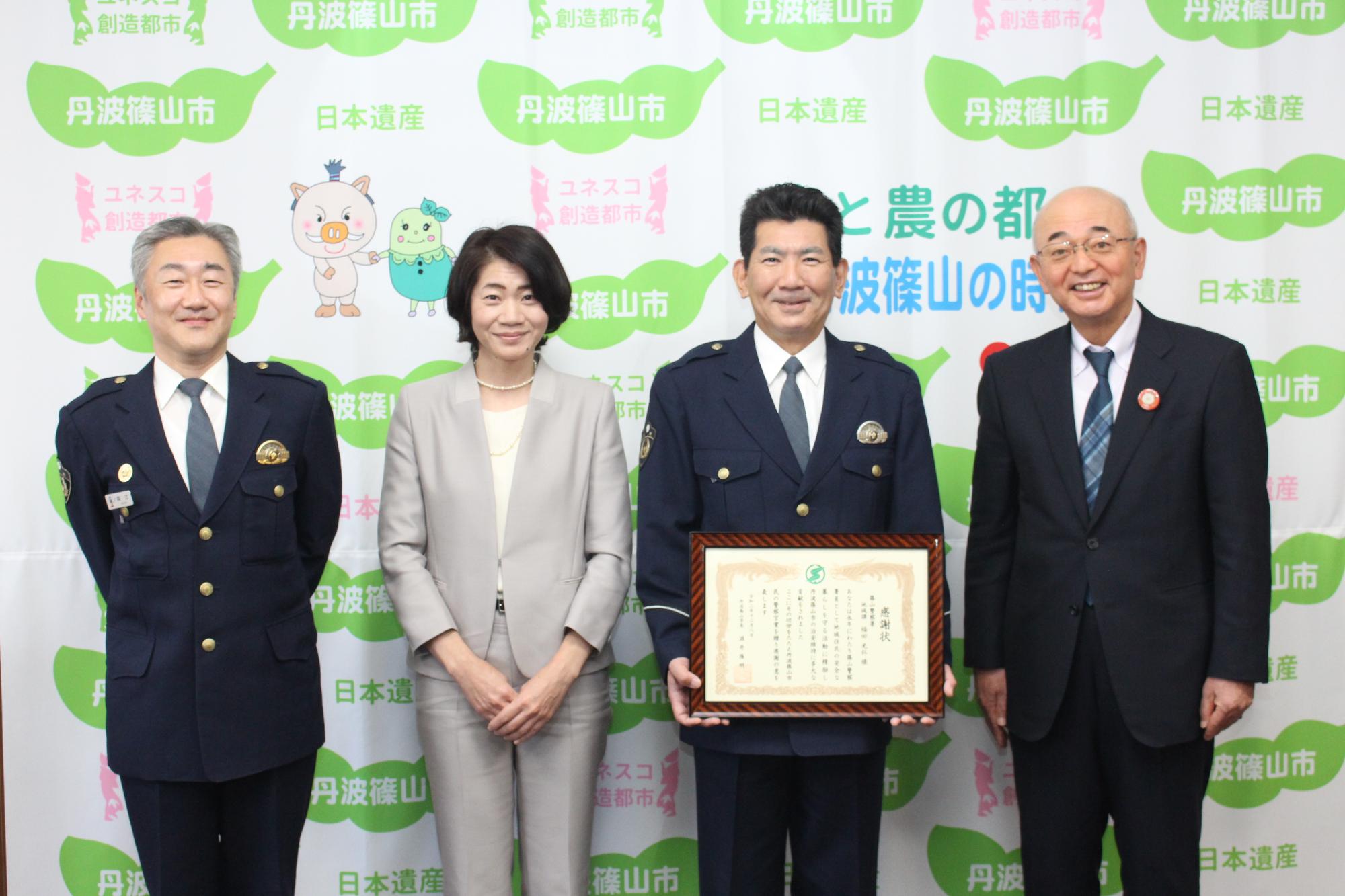 表彰状を手に持つ福田さんと奥様、その隣に市長と署長が並んで立っている