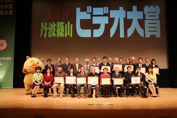 舞台のスクリーンに「第26回 丹波篠山 ビデオ大賞」と表示されており、その前に表彰された方が額縁に入った賞状を持っており、「まるいの」や観光大使の方や司会の熊谷さん、関係者が一緒に写っている集合写真