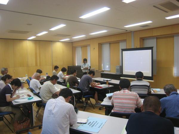 薄緑色の作業着を着ている女性が前に立って説明している講義を熱心に聞いている参加者の後ろ姿の写真