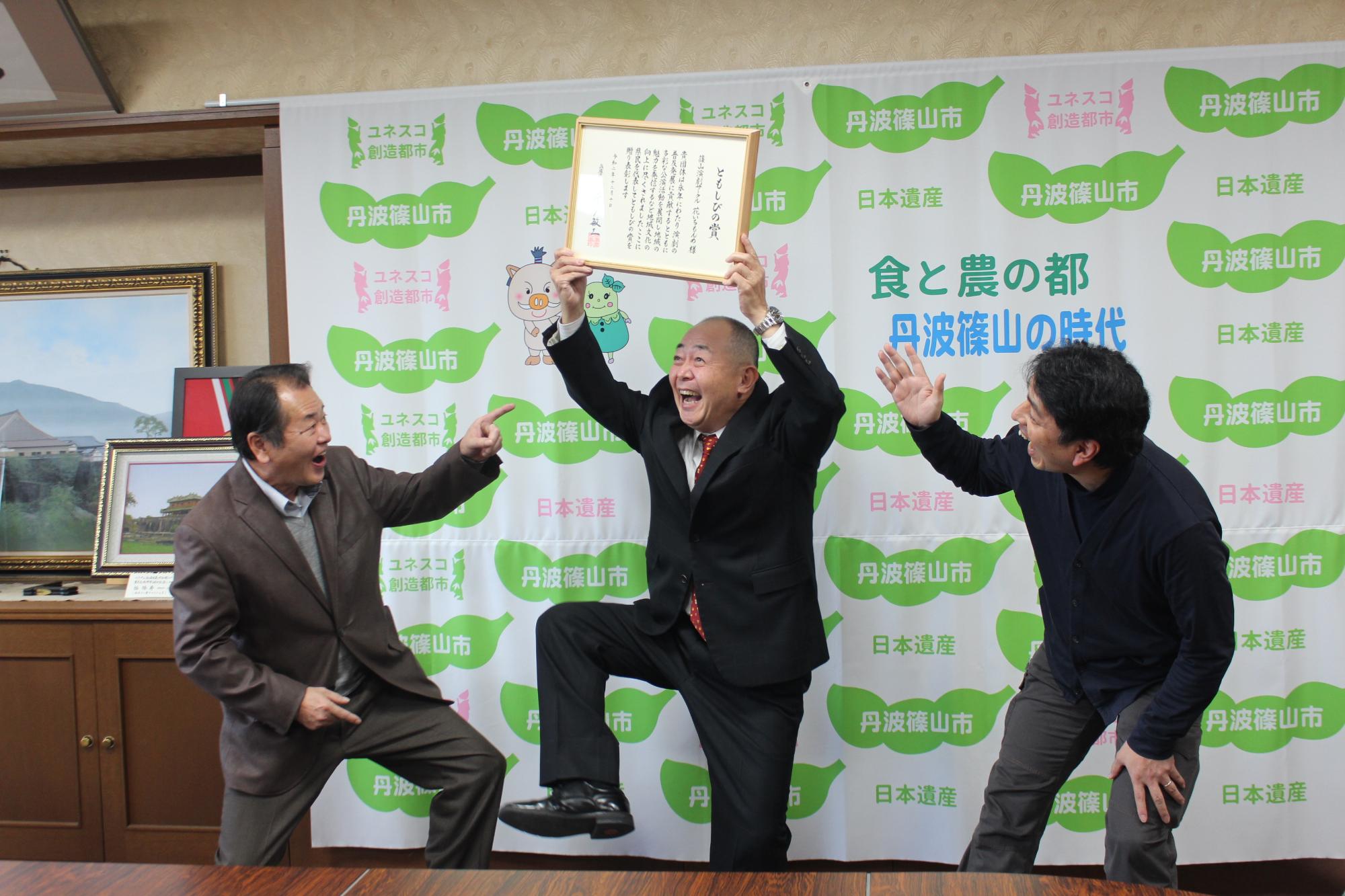 座長が表彰状を両手で持ち頭上にかかげ、片足あげて喜んでいる。団員二人が指をさしてびっくりしている。