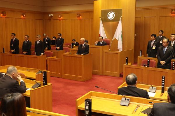 演壇に立っている酒井市長とその後ろに座っている議長、その両脇に立っている市職員と議員席に着席している議員の写真