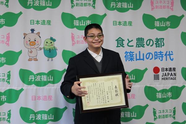 堂東 直矢さんが額に入った表彰状をもって笑顔で記念写真