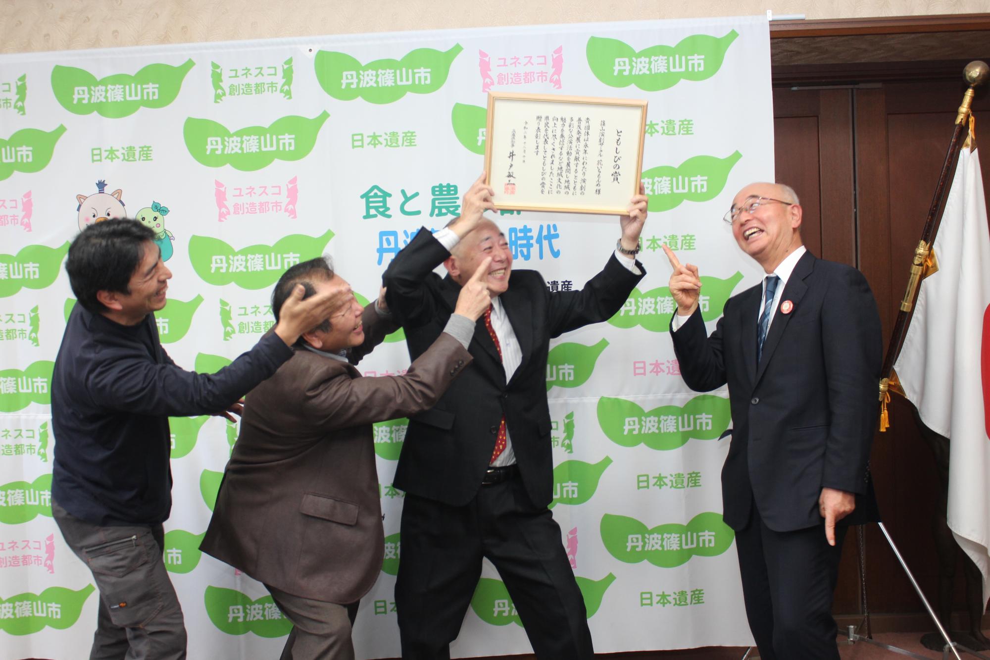 両手で持った表彰状を頭上にかかげ、団員二人と市長に表彰状を自慢する座長